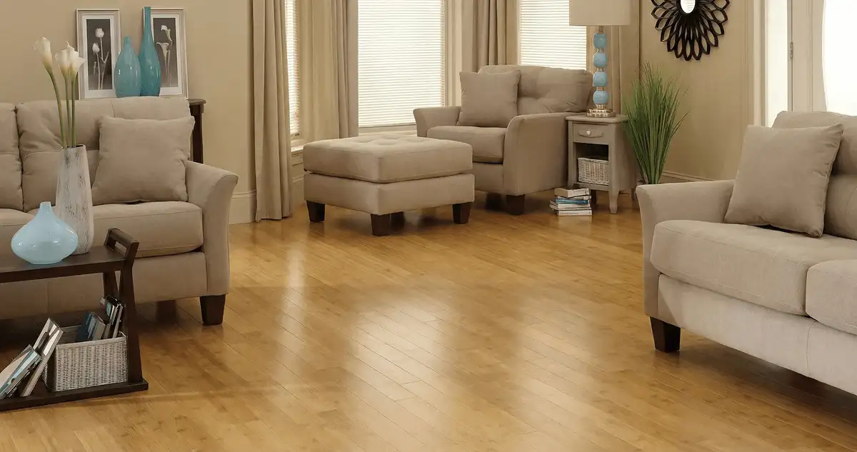 A living room with professionally installed bamboo flooring, beige sofas, a matching ottoman, a side table with a blue lamp, vases, framed pictures, and large windows with blinds and curtains in Mobile, AL