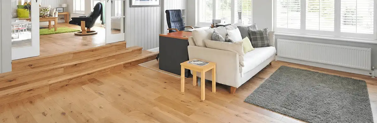 A modern living room with professionally installed laminate flooring, a beige sofa, a small side table, a gray rug, and large windows with white shutters in Mobile, AL